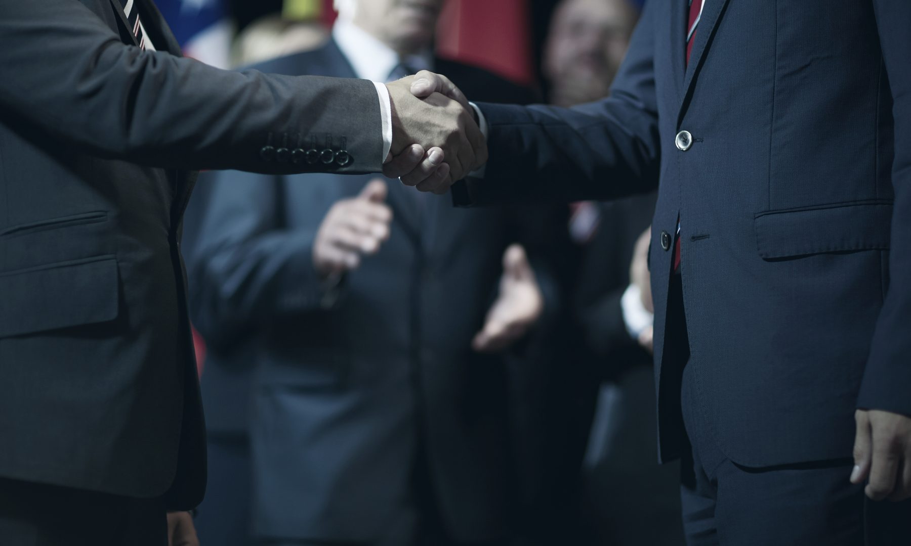 A candidate shaking hands with a supporter in front of flags at a campaign event